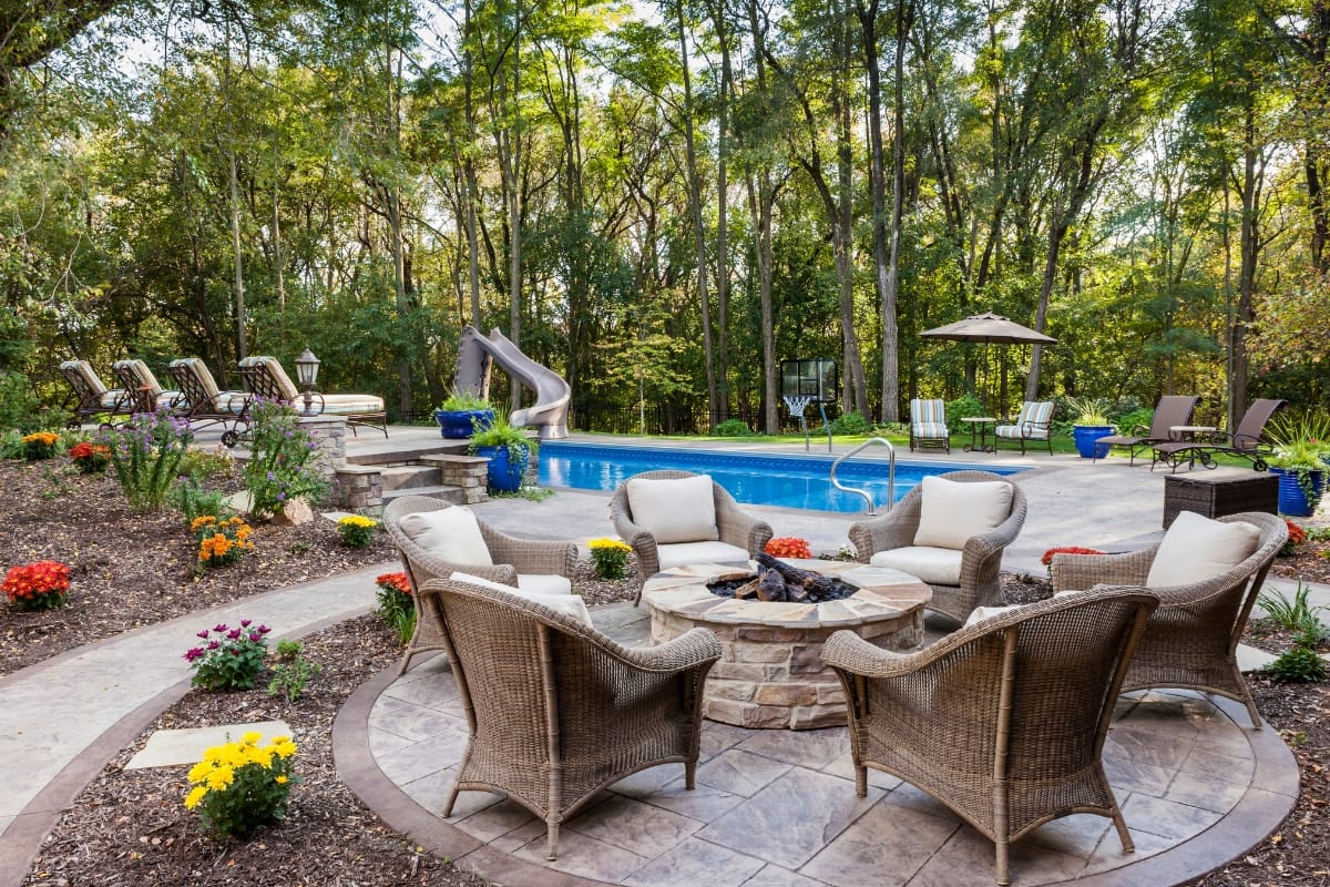 Backyard patio with wicker chairs around a cozy fire pit—perfect for trying out new fire pit ideas—a small pool with a slide, lounge chairs, and trees in the background.