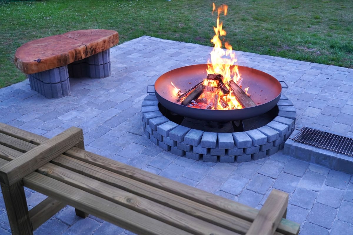 A round fire pit, serving as a centerpiece for your poolside charm, features burning logs surrounded by stone pavers. Nearby, a wooden bench and a seating area made of stone and wood offer inviting spaces to unwind on the patio under the glow of the flames.