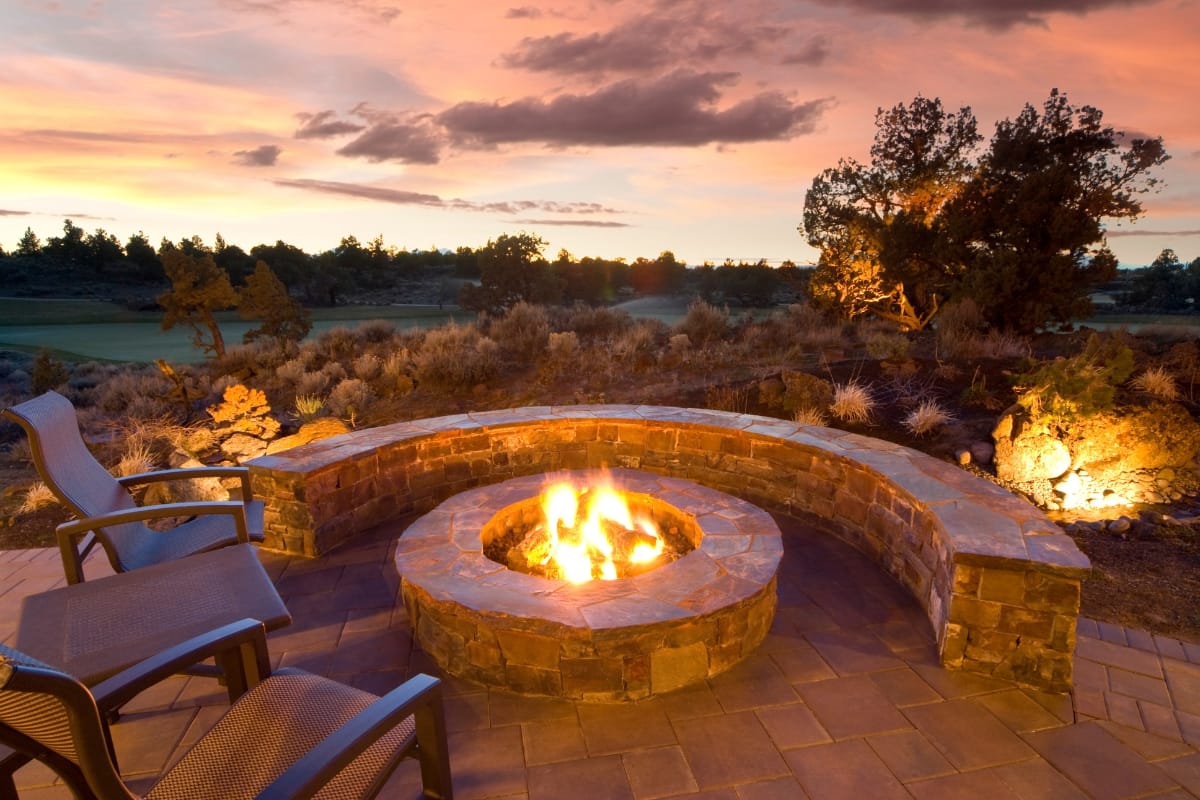 Curved stone bench encircles a lit fire pit on a patio with chairs, adding poolside charm against a vibrant sunset sky and natural landscape.