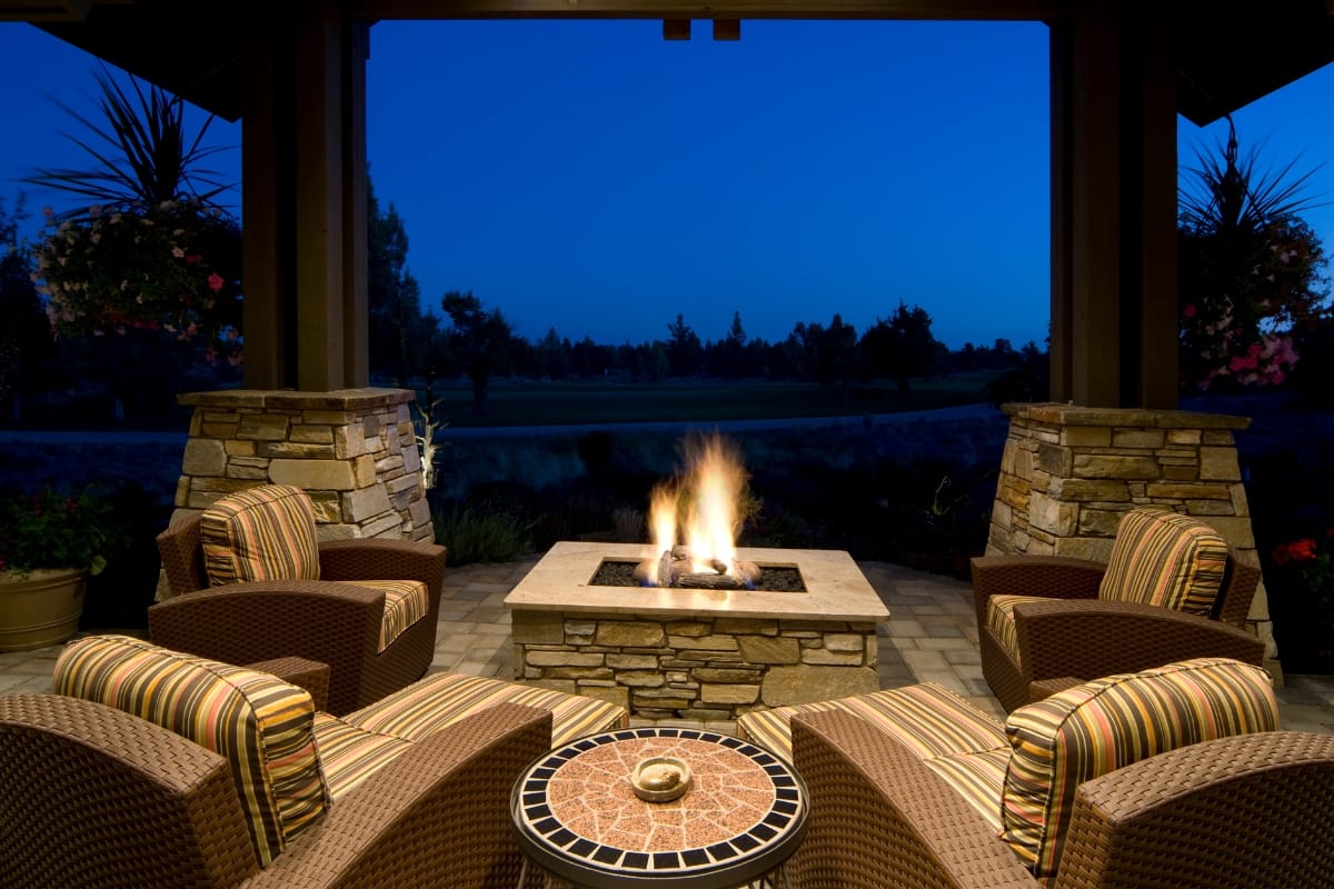 Under the covered patio, the outdoor seating area showcases poolside charm with a stone fire pit glowing warmly amidst four cushioned chairs. In this enchanting nighttime setting, the trees and sky provide a perfect backdrop for your fire pit ideas to ignite unforgettable evenings.