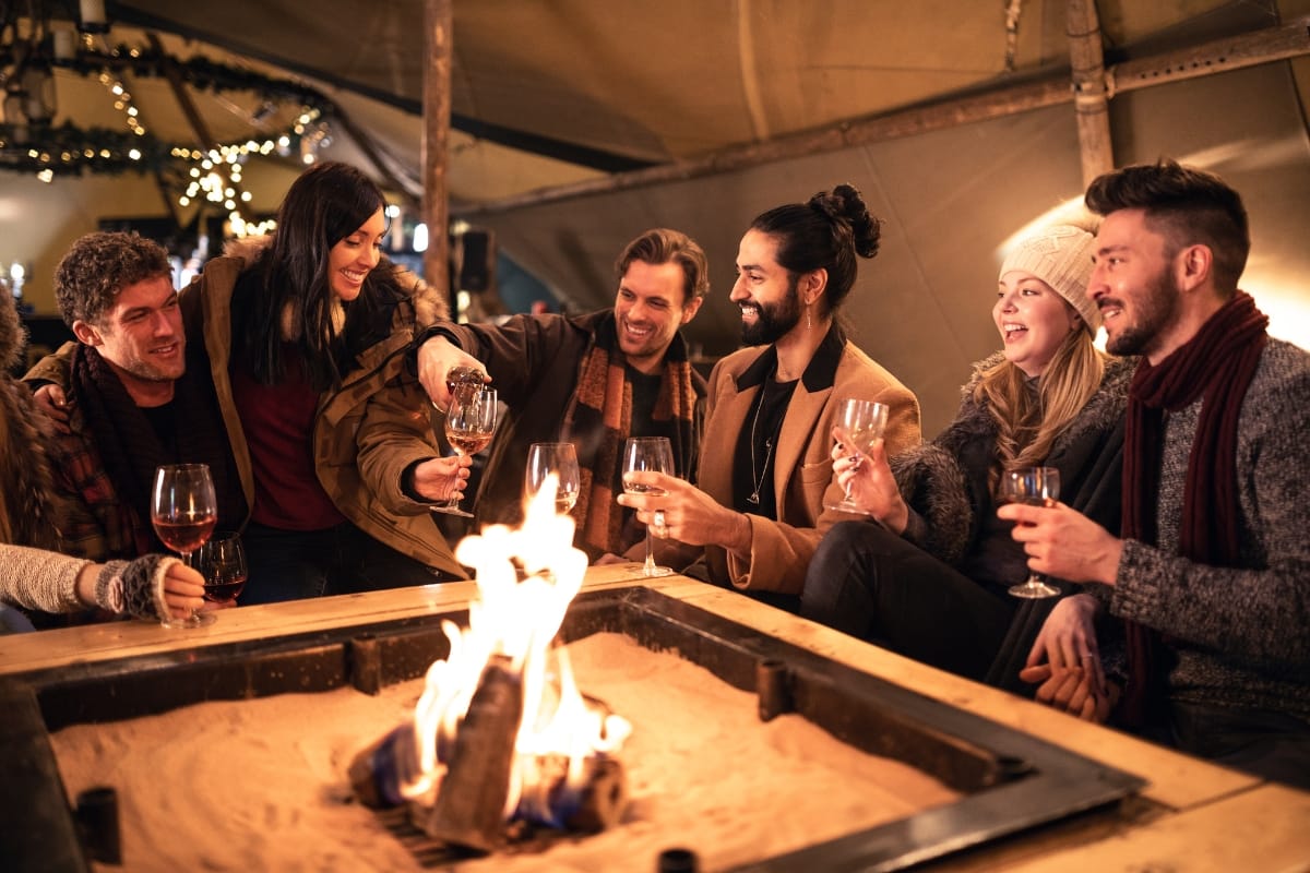 A charming group of people sit around a fire pit, toasting with wine glasses in a cozy setting.