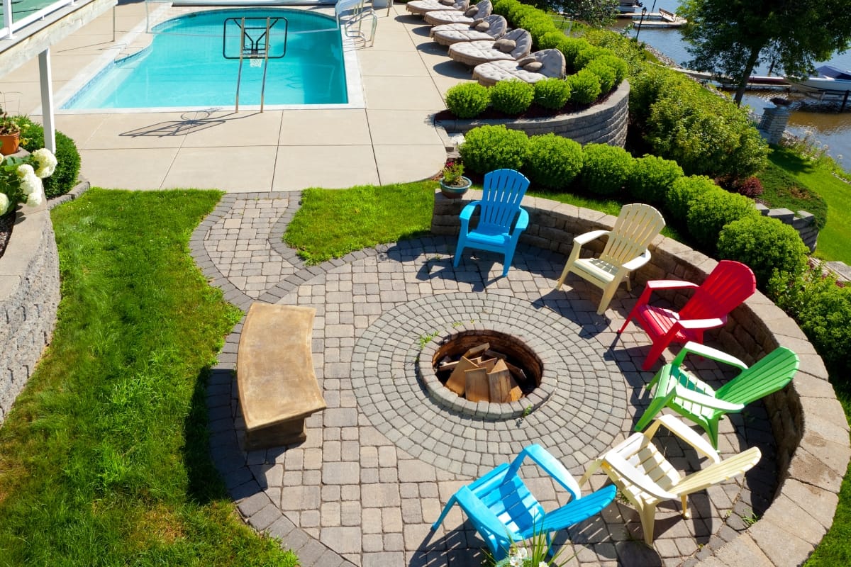 An inviting outdoor living space features a poolside fire pit surrounded by colorful chairs on a patio amidst lush greenery, with a serene body of water visible in the background.