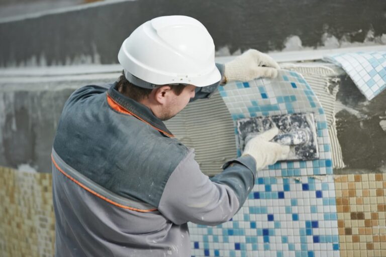 A construction worker in a hard hat installs blue and beige mosaic tiles on a wall, using adhesive and a trowel—a meticulous process that can impact overall pool renovation costs.