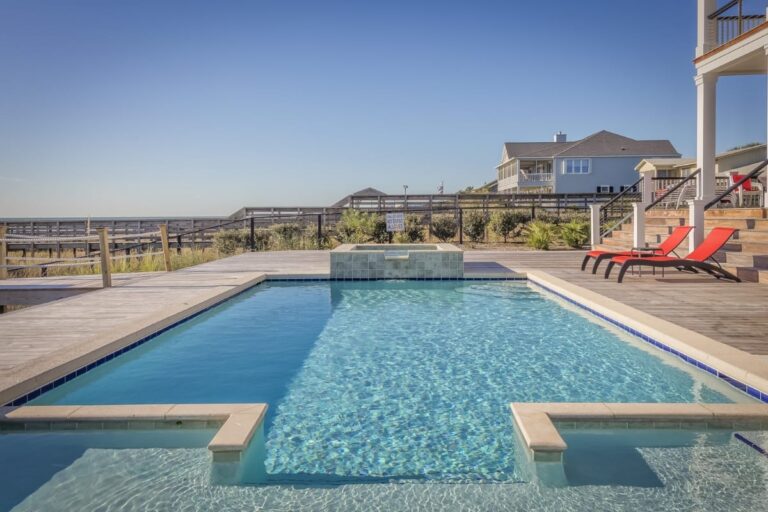 Outdoor swimming pool with clear blue water, recently refreshed through a pool renovation process, surrounded by a wooden deck, red lounge chairs, and a coastal house in the background under a clear sky.