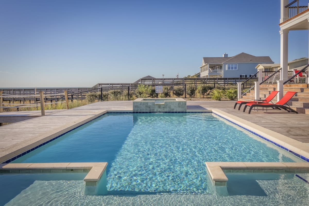 Outdoor swimming pool with clear blue water, recently refreshed through a pool renovation process, surrounded by a wooden deck, red lounge chairs, and a coastal house in the background under a clear sky.