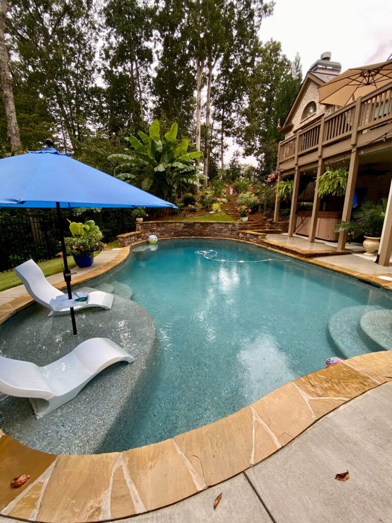 A backyard swimming pool with two lounge chairs under a blue umbrella, surrounded by plants, a stone patio, and a wooden deck, indicating a well-kept area through regular pool maintenance.