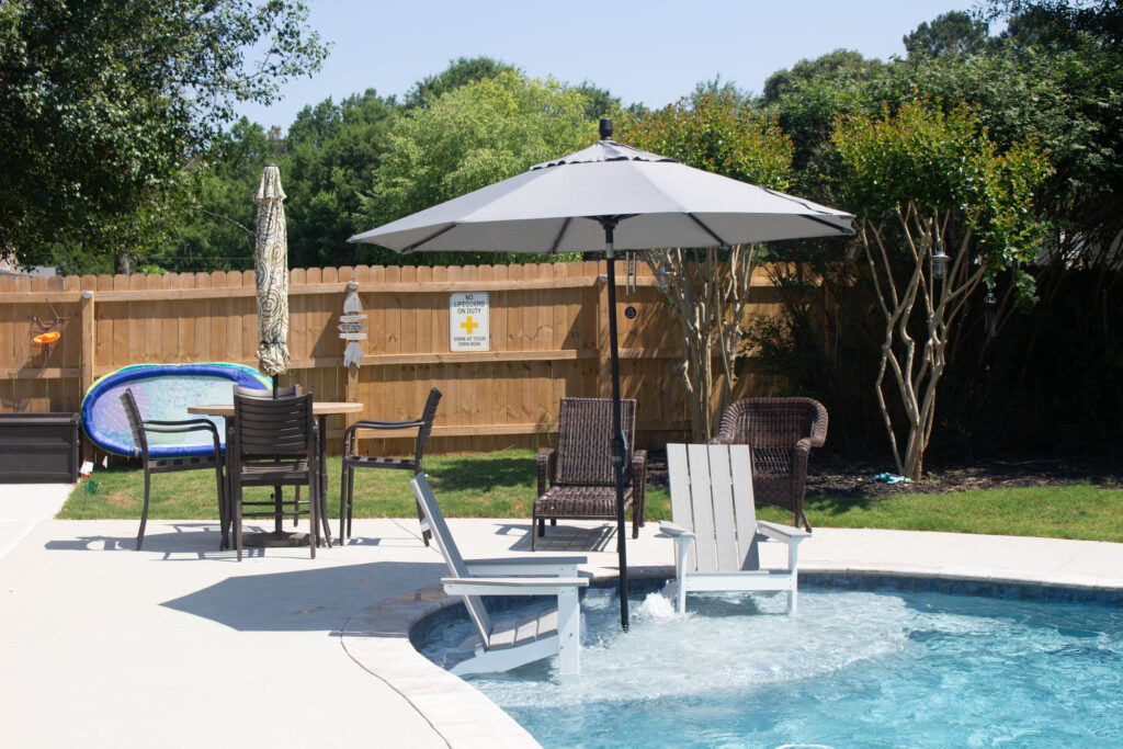 Two chairs with an umbrella are placed in the shallow end of a pool, with patio furniture and a wooden fence in the background.