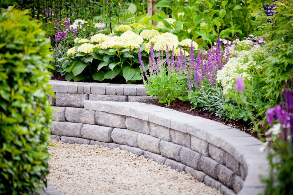 Curved custom retaining walls borders a gravel path, with green shrubs and blooming flowers in shades of white, yellow, and purple planted above the wall.