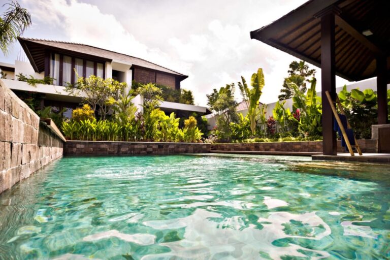 A residential backyard features a clear swimming pool with recent pool resurfacing, lush tropical plants, and a modern house with large windows and a patio.
