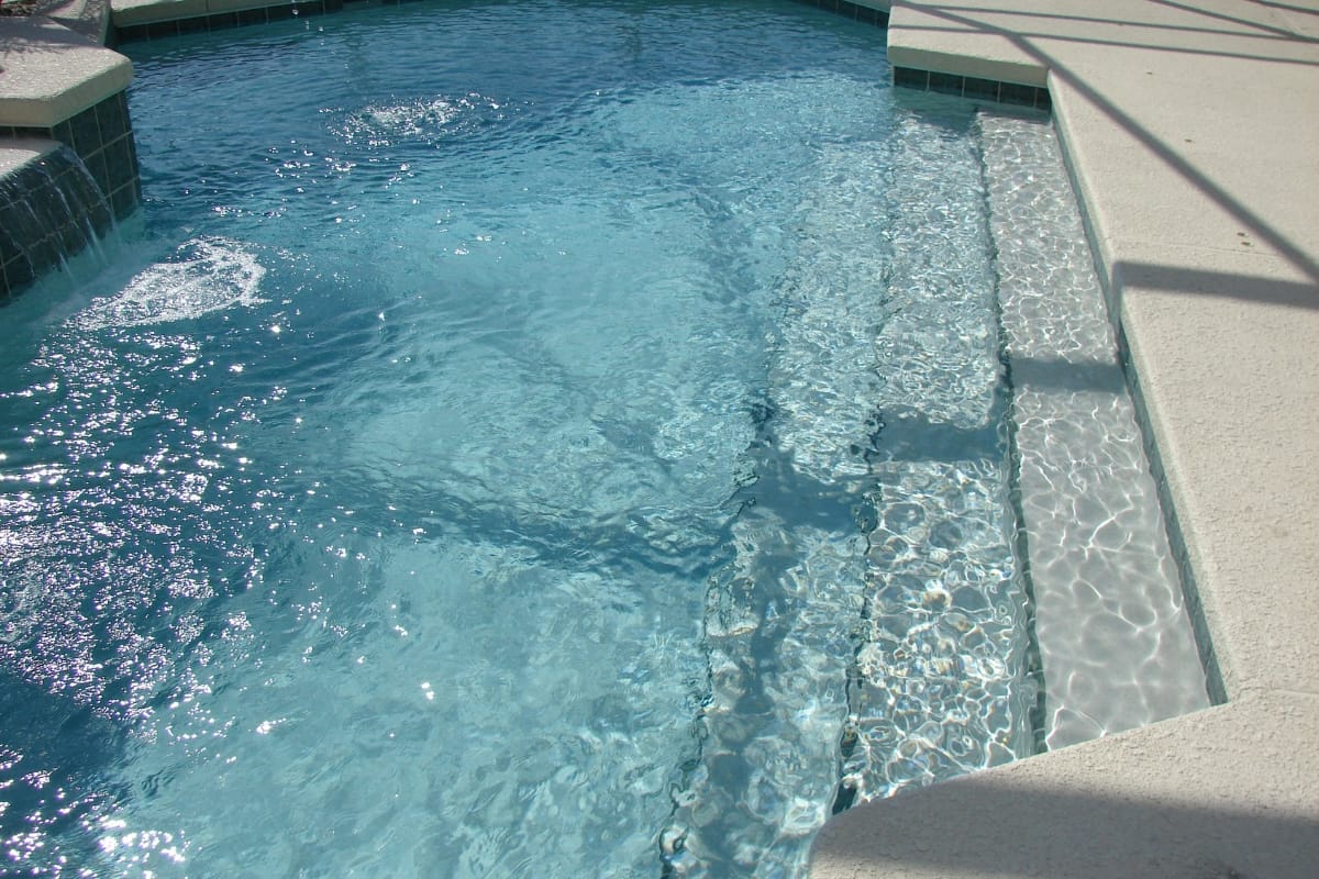 Sunlit swimming pool with clear water, featuring concrete steps leading in and a tiled edge—ideal for showcasing recent pool resurfacing.