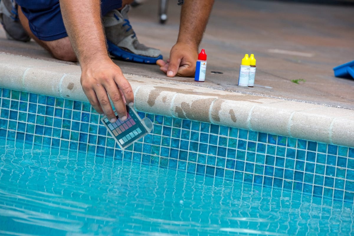 A person holds a water testing strip at the edge of a swimming pool, with pool chemical test kits placed nearby on the tile—a key step in maintaining water quality after pool resurfacing.