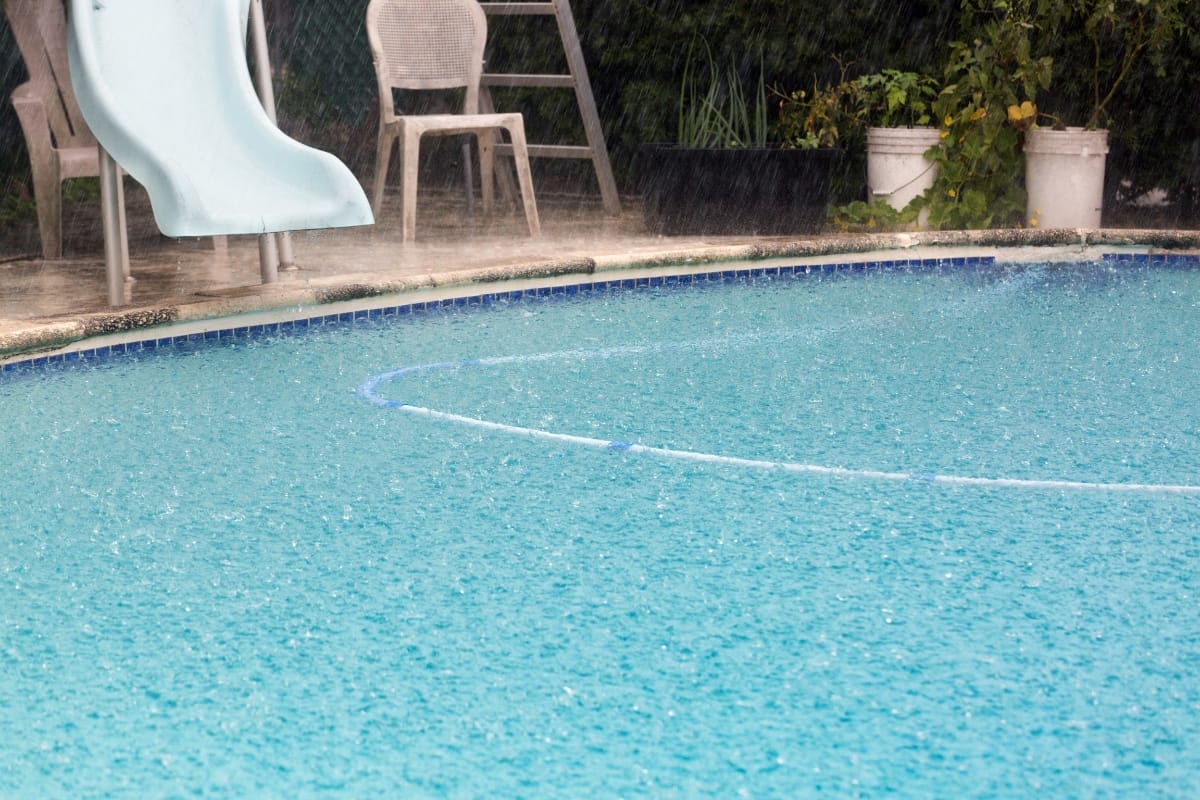 A swimming pool is shown during rainfall, with visible raindrops on the water. Pool resurfacing may be needed for maintenance, as a slide, chairs, and white buckets are seen near the poolside.