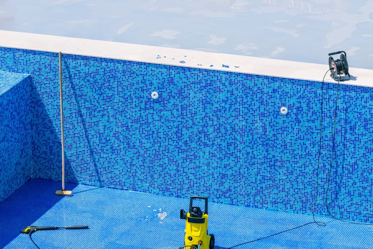 Empty swimming pool with blue mosaic tiles, cleaning tools, a broom, and a pressure washer inside—ideal for pool resurfacing. A hose reel sits at the pool edge, highlighting preparation for restoration work.