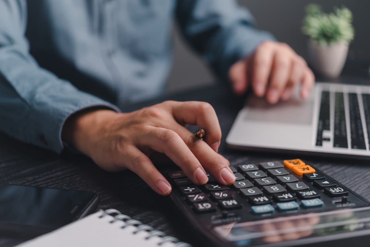 Person using a calculator and laptop on a desk with a notepad, pen, and a small potted plant nearby, possibly calculating expenses or planning for pool resurfacing.
