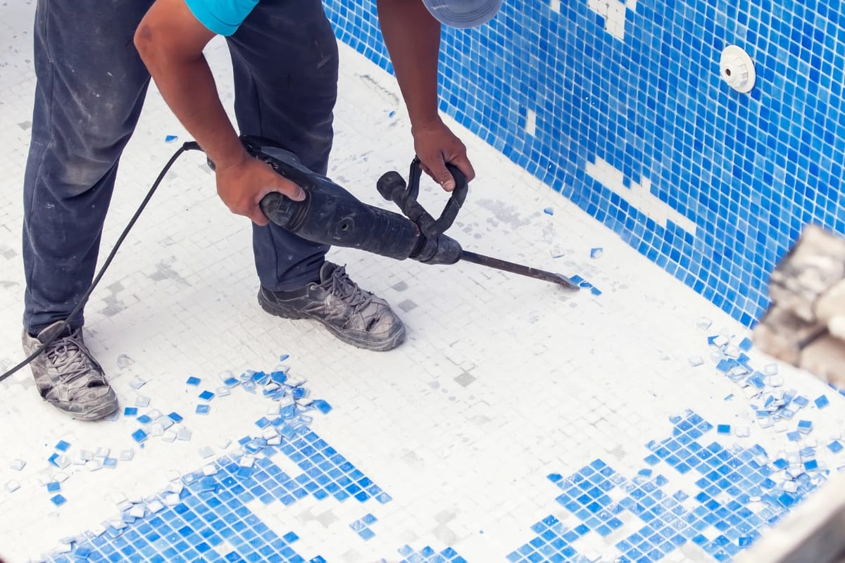 A person uses a power tool for pool resurfacing, removing blue and white tiles from the bottom of an empty swimming pool. Broken tiles are scattered on the floor.