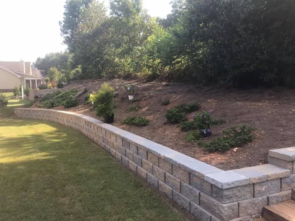 A retaining wall made of stone blocks separates a grassy lawn from a sloped garden area with small shrubs and young plants.