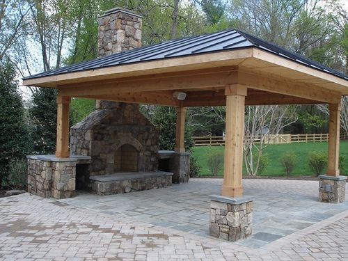 A covered outdoor patio with a stone fireplace, wooden beams, and a sloped roof, set on a paved area surrounded by greenery.