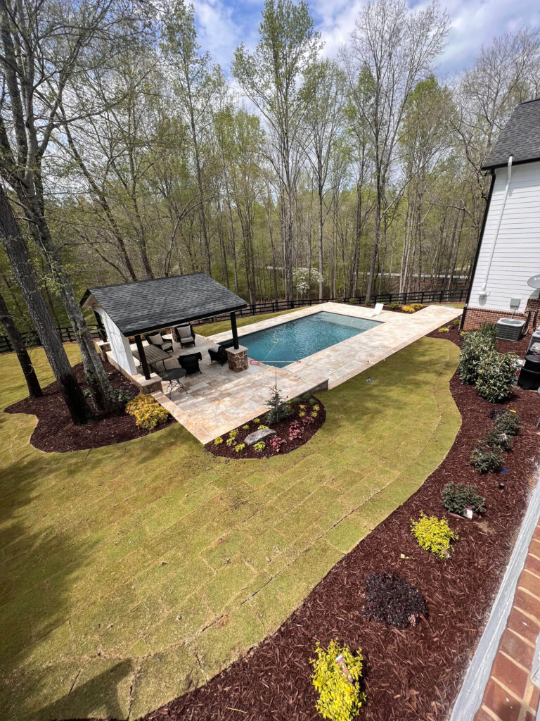 A rectangular backyard swimming pool with a stone patio, covered seating area, and newly landscaped yard bordered by trees.