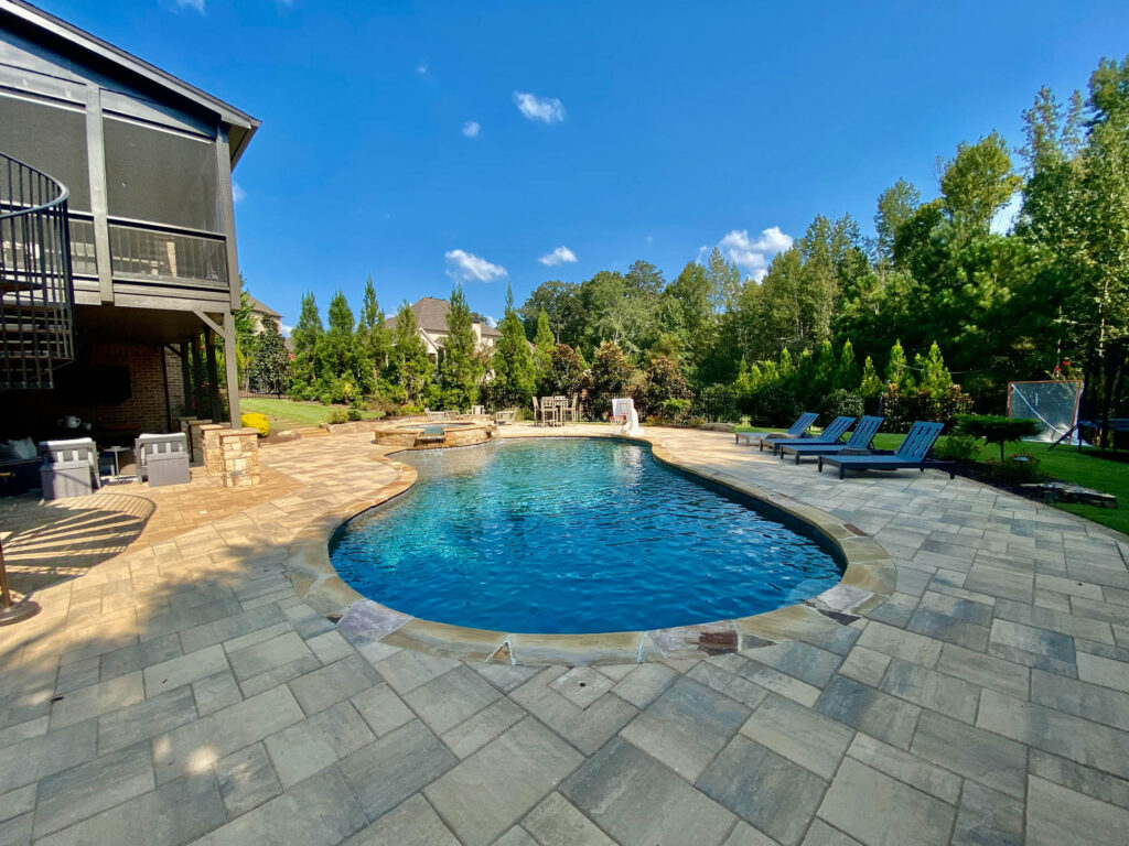 A backyard swimming pool with lounge chairs, a hot tub, and landscaping; a two-story house with a spiral staircase is visible on the left, showcasing the potential for incredible pool remodeling.