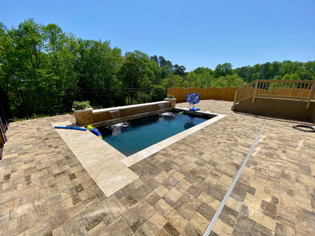 Rectangular backyard pool surrounded by tan stone pavers, with pool toys floating in the water and trees in the background.