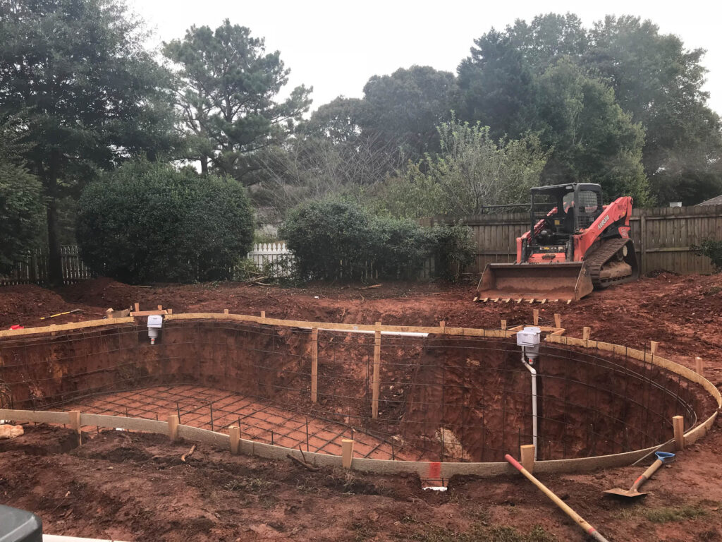 Construction site with an in-ground pool being built; rebar and wooden framing are visible, and a bulldozer is parked on the dirt nearby.