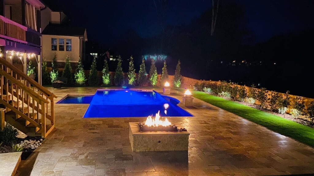 A backyard at night featuring a lit fire pit, a glowing blue swimming pool, manicured lawn, and outdoor lighting along the fence—a luxurious scene often brought to life by expert pool builders in Dawson County.
