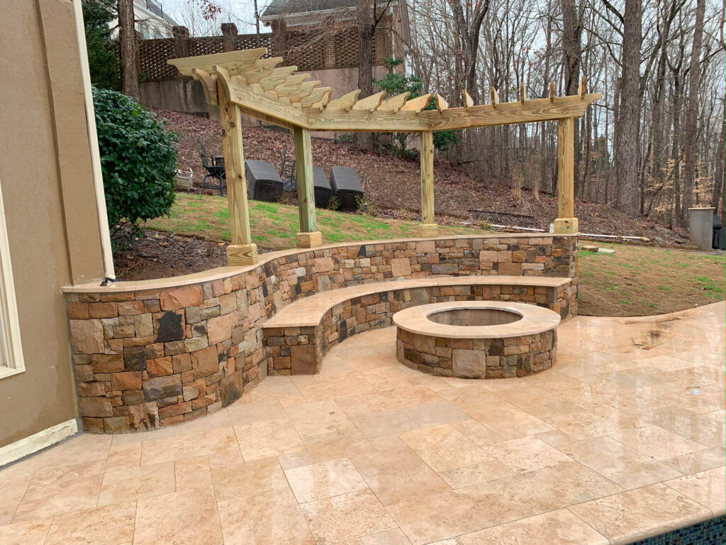 Stone patio with a curved stone bench, round fire pit, and wooden pergola, set against a wooded backyard.