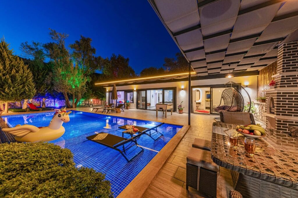 Modern backyard at dusk with a lit swimming pool featuring some of the best pool accessories like a stylish pool float, lounge chairs, patio seating, and a covered outdoor dining area with drinks and fresh fruit on the table.