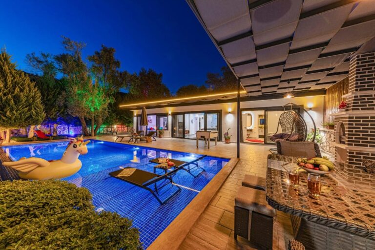 Modern backyard at dusk with a lit swimming pool featuring some of the best pool accessories like a stylish pool float, lounge chairs, patio seating, and a covered outdoor dining area with drinks and fresh fruit on the table.