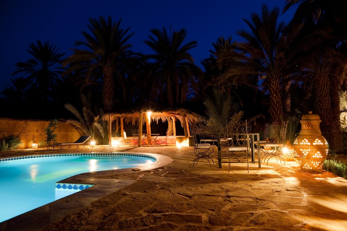 A swimming pool with glowing water is surrounded by stone tiles, outdoor furniture, and palm trees, featuring the best pool accessories and illuminated by warm lights at night.