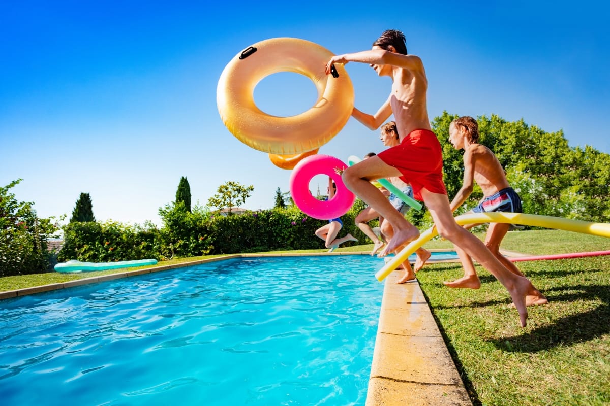 Four people with the best pool accessories—colorful inflatable rings—jump into an outdoor swimming pool on a sunny day, surrounded by lush greenery.