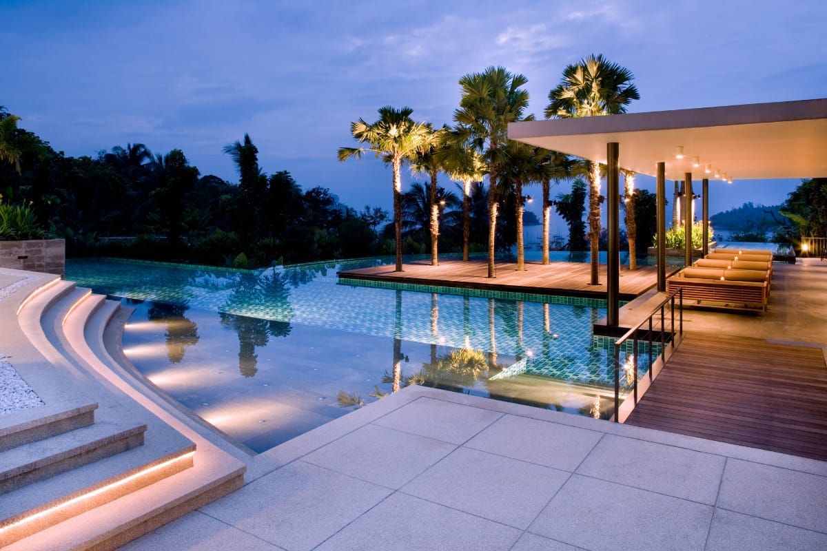 Modern outdoor pool area at dusk with palm trees, illuminated steps, lounge chairs under a canopy, and reflections in the water—elevate your experience with the best pool accessories for comfort and style.