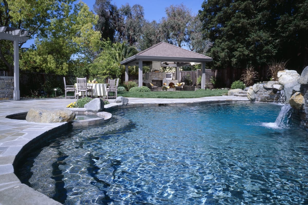 A backyard swimming pool with a rock waterfall, gazebo, patio seating area, green lawn, and surrounding trees on a sunny day—perfect for relaxing or enjoying the best pool accessories.