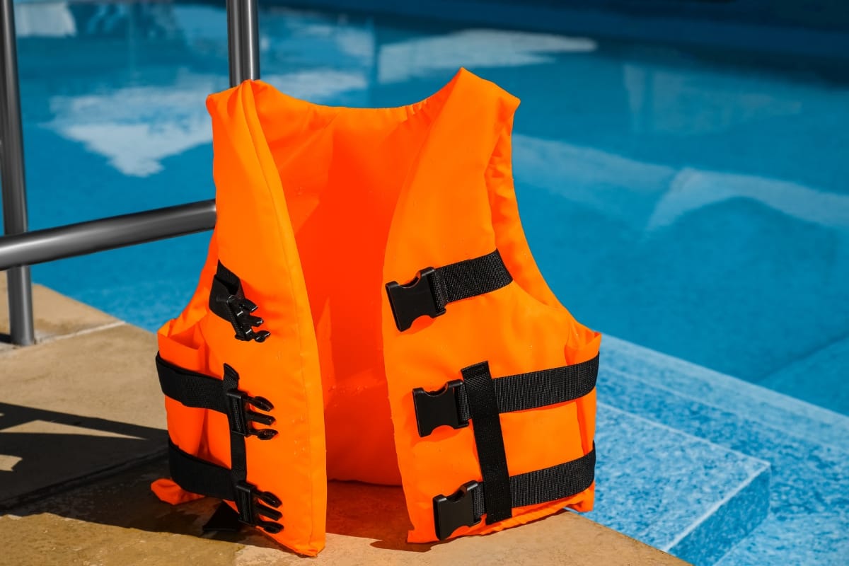 An orange life jacket with black straps, one of the best pool accessories, is placed on the poolside next to a swimming pool.