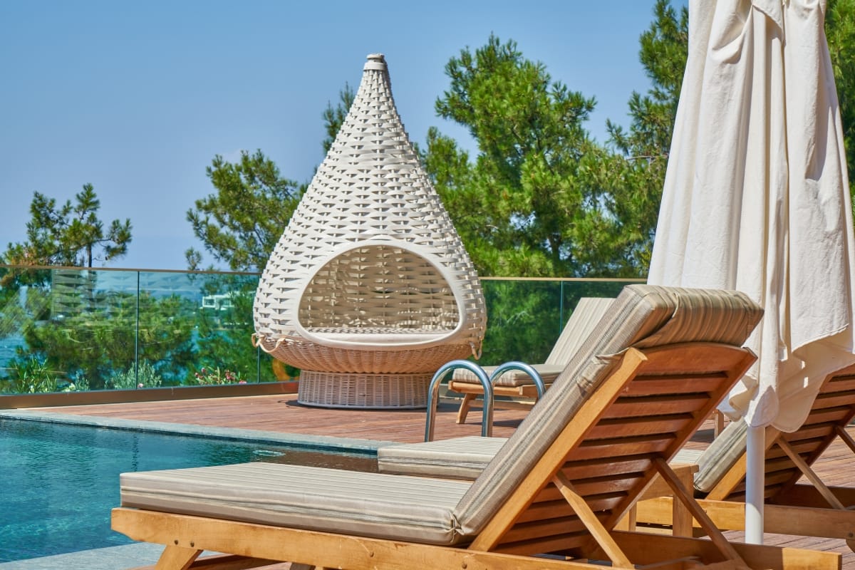 Poolside scene with wooden lounge chairs, a white wicker egg-shaped chair, an umbrella, and trees in the background on a sunny day, featuring some of the best pool accessories for relaxing in style.