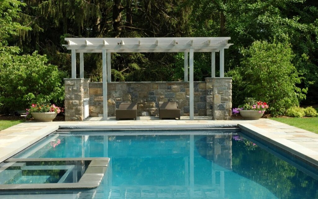 Rectangular swimming pool with a pergola and lounge chairs in the background, surrounded by greenery and potted flowers.