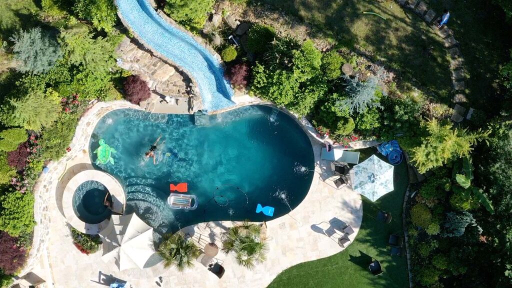 Inground Swimming pool construction in Atlanta: Aerial view of a backyard pool with a waterslide, hot tub, pool floats, lounge chairs, umbrella, and surrounding landscaping.