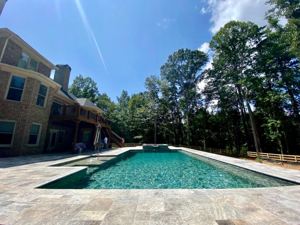 Pool Financing in Atlanta: Rectangular outdoor swimming pool with clear water next to a large brick house, surrounded by trees and a stone patio under a sunny sky.