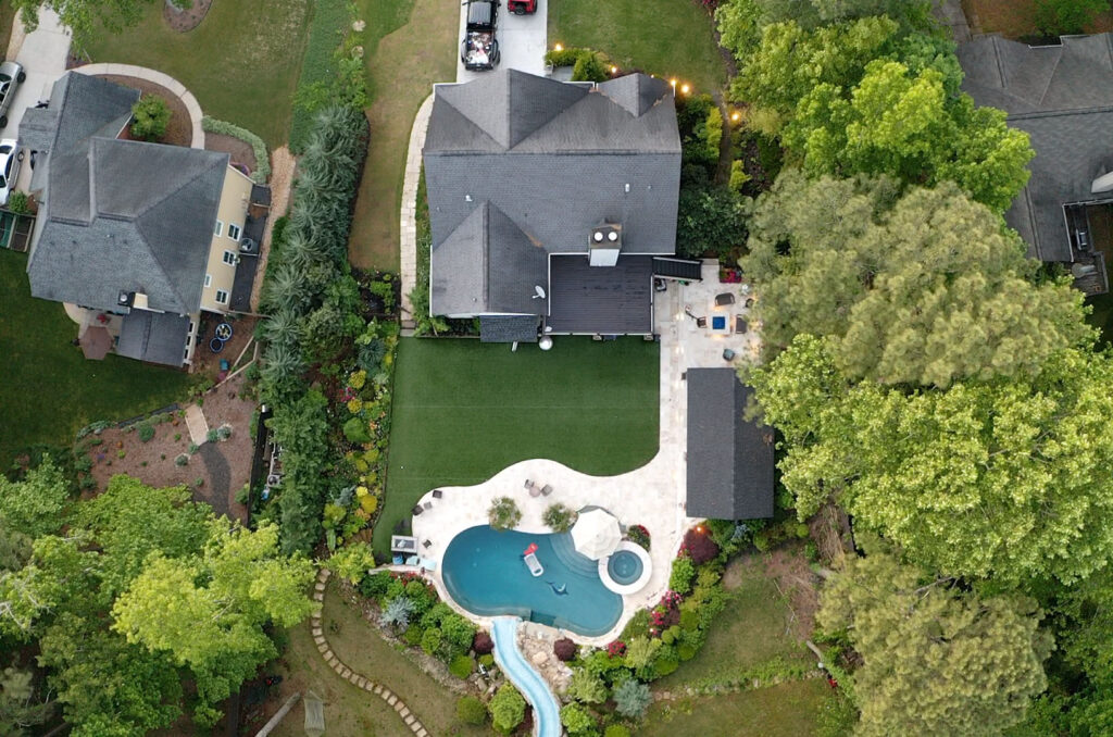 Aerial view of a house with a backyard featuring a swimming pool, water slide, patio area, and surrounding trees and landscaping.