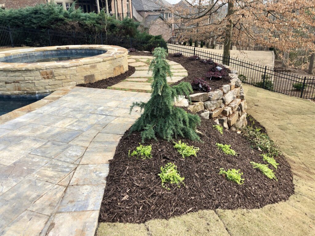 This landscaped backyard showcases detailed retaining walls and stonework through its stone pathway and circular stone hot tub. The garden also includes a mulched plant bed, small shrubs, and a young conifer tree, creating a cohesive outdoor space.