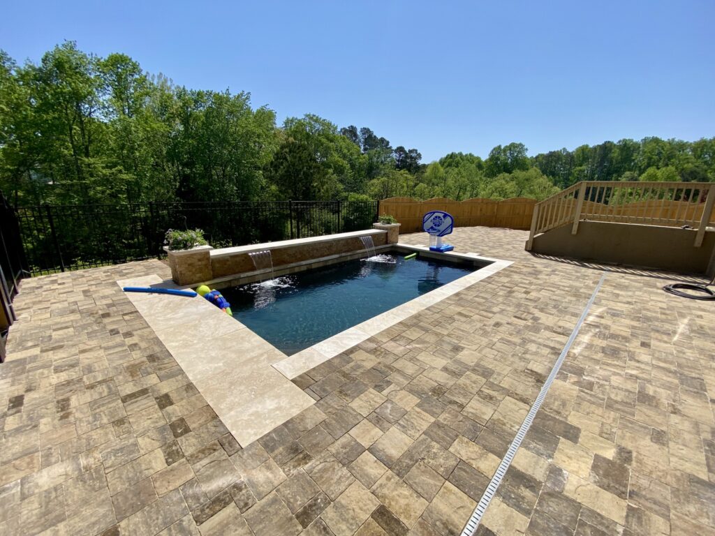 Rectangular backyard pool with toys, surrounded by a paved patio and fenced by trees, under a clear blue sky. A wooden deck is visible on the right side.