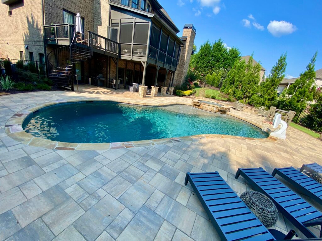 A vibrant backyard featuring an in-ground swimming pool with a small slide, a stone patio, and lounge chairs. Check our pool specials to create a similar oasis, all set against a two-story brick house.