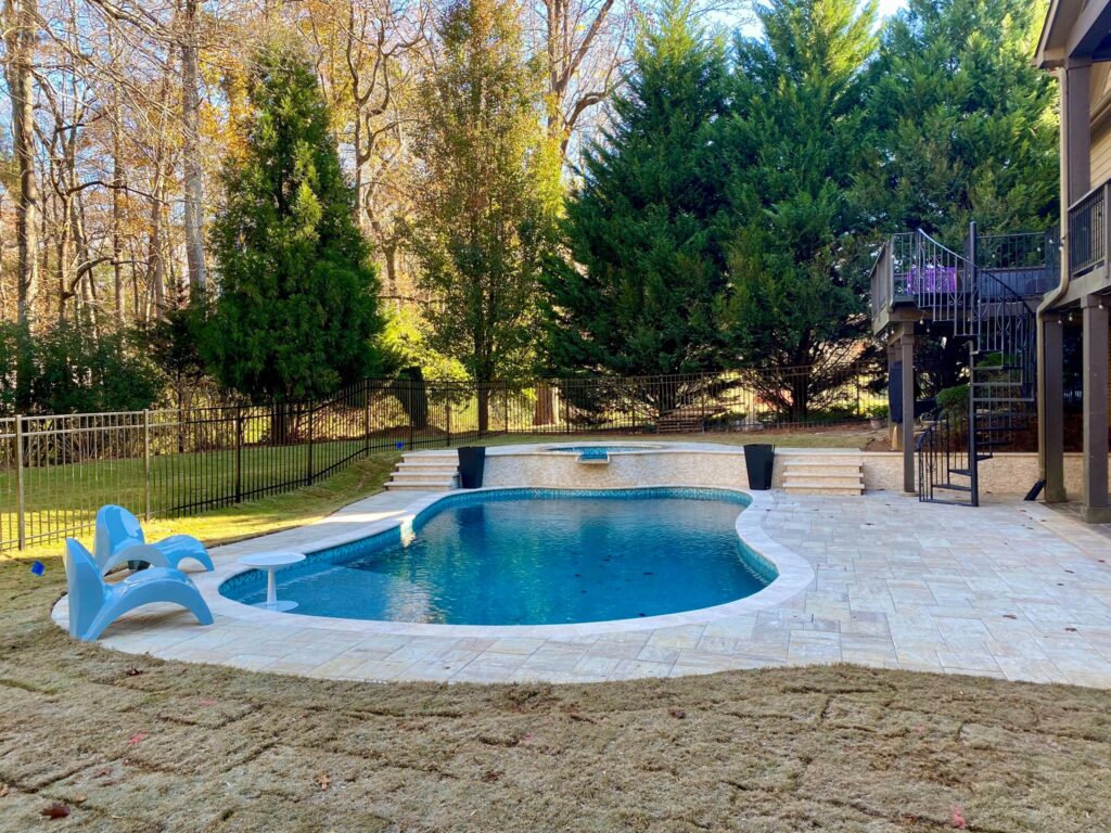 Backyard swimming pool with white tile deck, pool steps, two blue lounge chairs, black fence, and surrounding trees. A spiral staircase leads to an upper deck on the right—contact Clear Water Pools to create a custom design like this.