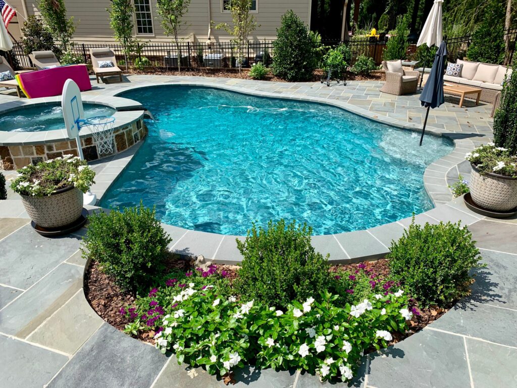 A backyard pool with clear blue water, a connected spa, stone patio, potted plants, and outdoor seating area with cushions and umbrellas, ready for enjoyment thanks to professional pool repair in Atlanta.