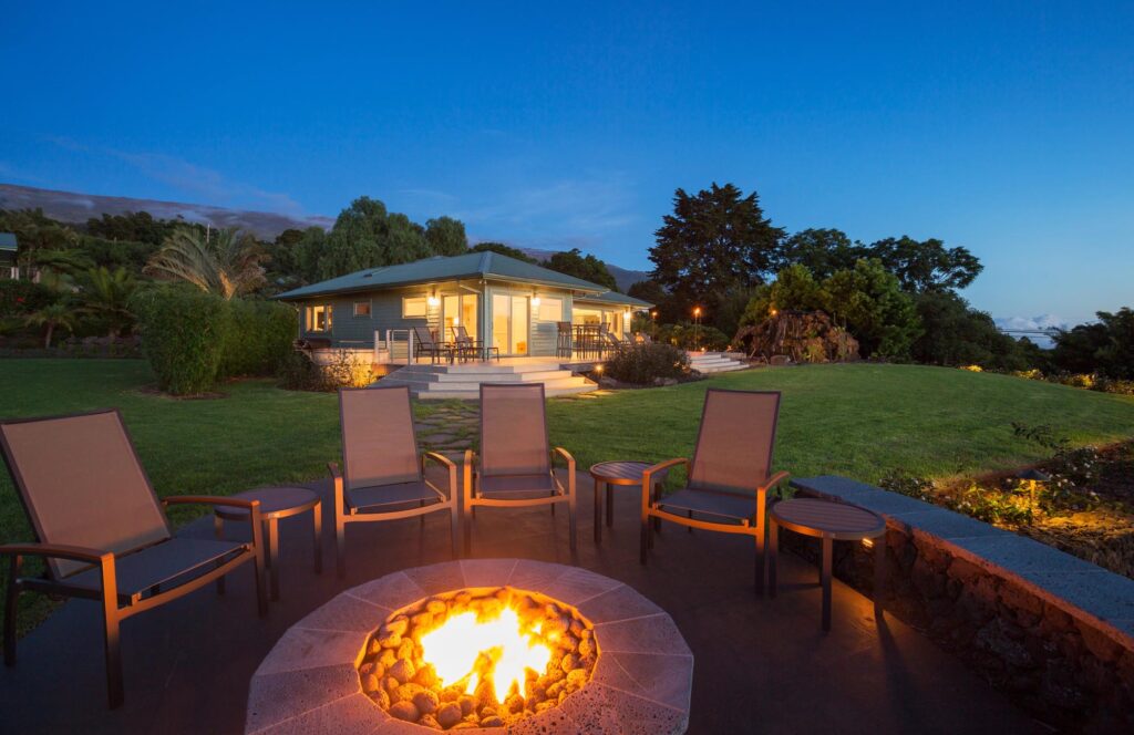 A modern house with a large lawn is seen at dusk, with five chairs arranged around a lit fire pit in the foreground.