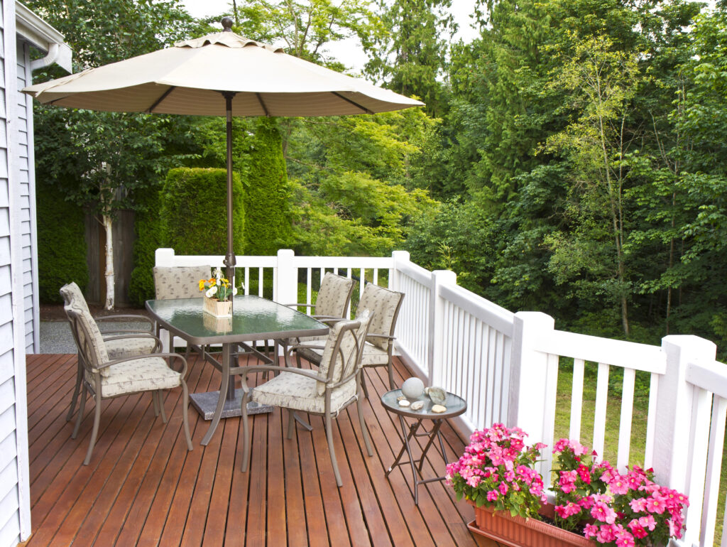 A patio with a glass table, six cushioned chairs, and a large umbrella sits on a wooden deck surrounded by white railing, flowers, green trees, and expert landscape design.