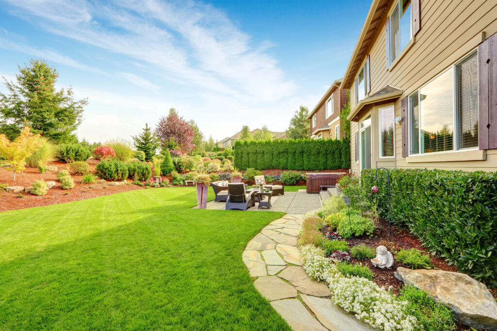 Backyard with green lawn, professional landscape design, landscaped garden beds, patio furniture, and a stone walkway beside a house on a sunny day.