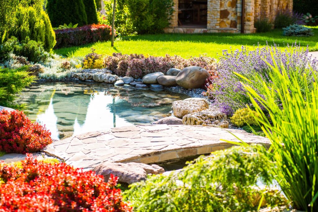 A landscaped garden with a small pond, stone bridge, various plants, and shrubs under bright sunlight, demonstrating exceptional landscape design installation.