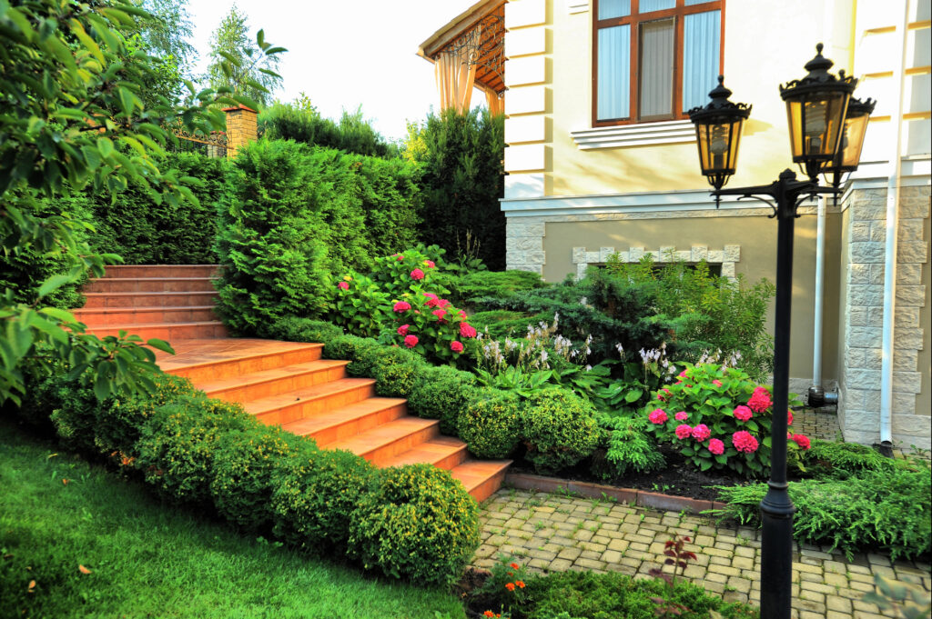 A landscaped garden with expert landscape design features terracotta steps, manicured shrubs, blooming pink flowers, a stone path, and a vintage-style lamp post beside a beige house.