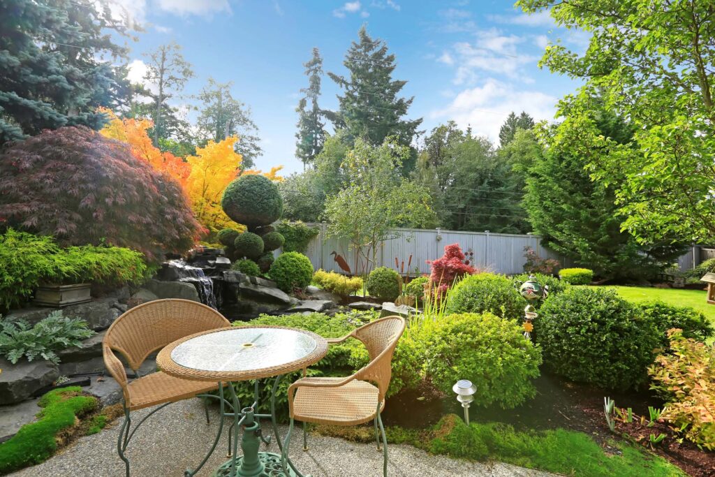 A round glass-top table with two wicker chairs sits on a patio in a landscaped garden with lush greenery, a small waterfall, and bright autumn foliage.